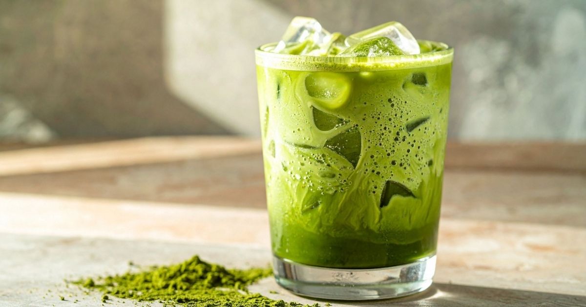 Vibrant green matcha latte in a clear glass next to a bamboo whisk and bowl of matcha powder